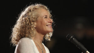 Carole King smiling sitting at piano microphone