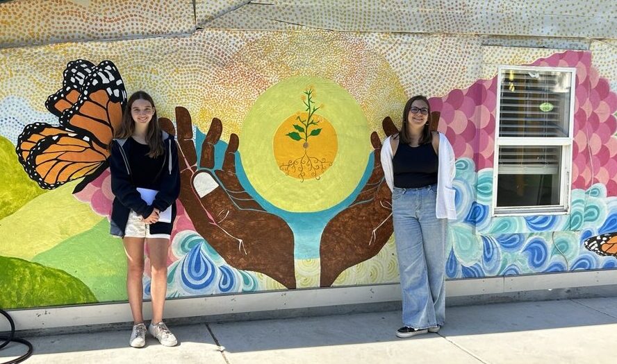 Lily and Avery standing in front of Mustard Seed School mural